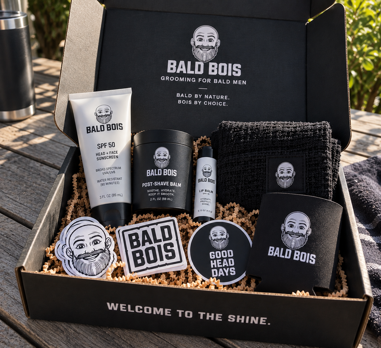 A Bald Bois box with SPF head sunscreen, post-shave balm, lip balm, a towel, stickers, and a koozie